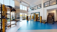 Located at 7200 N 91st Ave in Glendale, Arizona, this fitness center offers a generously sized, single-floor exercise space designed to accommodate a wide range of workout needs. The facility is defined by its dramatic double-height ceilings, which create an open, airy atmosphere and are complemented by an upper-level mezzanine accessible via a modern staircase with a sleek navy blue metal railing and cable detailing.

Large floor-to-ceiling windows line the perimeter, flooding the interior with abundant natural light and providing views of the surrounding outdoor areas. The flooring features a distinctive two-tone design, combining crisp white and vibrant blue rubber surfaces that delineate different workout zones throughout the space.

The main floor is comprehensively equipped with a robust selection of yellow-accented strength machines, including cable systems, multi-functional training rigs, plate-loaded machines, and free weight racks with benches. Full-length framed mirrors with navy trim are strategically positioned along the walls to enhance spatial perception and assist users during exercise. The mezzanine level houses additional cardio equipment, providing an elevated vantage point over the main floor. The overall design is modern and functional, with white walls, exposed ductwork, and recessed lighting contributing to a clean, professional environment.
