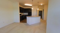 This single-story space showcases an open-concept layout that connects the kitchen and living areas in a cohesive, functional design. The kitchen is a standout feature, outfitted with rich, dark espresso cabinetry that extends to the upper and lower walls, providing generous storage. Black appliances, including a refrigerator, range, and built-in microwave, complement the bold cabinetry for a sleek, uniform aesthetic. A central kitchen island with a light-toned countertop and an integrated sink serves as the focal point, illuminated by two elegant pendant lights that hang from above, adding both task lighting and visual interest. The transition between the kitchen's hardwood-style flooring and the plush carpeting of the adjacent living area subtly defines each zone while maintaining an open feel. A graceful arched doorway leads to a hallway beyond, adding an architectural detail that softens the overall design. Neutral wall tones throughout create a clean, inviting backdrop that complements the contrasting dark finishes. The layout offers a practical and stylish environment well-suited for both everyday living and entertaining.