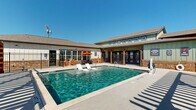 Located at 1666 W Cumberland Rd in Tyler, Texas, this outdoor amenity area is centered around a generously sized resort-style swimming pool with crystal-clear water. The pool features built-in in-water lounge chairs with water jets, providing a relaxing wading experience, as well as traditional poolside chaise lounges for sunbathing on the expansive concrete deck. The pool area is clearly marked with depth indicators and no-diving signage, reflecting a well-maintained and safety-conscious facility. Two substantial wooden pergola structures flank the pool area, offering shaded retreats furnished with additional lounge chairs and seating. Colorful Adirondack-style chairs and umbrella-shaded tables are arranged around the deck, adding a vibrant and inviting atmosphere. A single-story clubhouse building borders one side of the pool, featuring a combination of board-and-batten siding, brick accents, and a pitched shingle roof. The building includes large multi-pane windows and French-style double doors that open directly onto the pool deck, creating a seamless indoor-outdoor connection. A covered outdoor corridor with a timber-framed pergola extends from the clubhouse, adding architectural character. The entire pool area is enclosed by a black iron fence, ensuring security while maintaining open sightlines to the surrounding Texas sky.