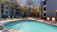 This inviting outdoor pool area serves as a central amenity within a multi-story residential community. The generously sized swimming pool features clear, blue water and is complemented by an adjacent spa, offering a range of relaxation options for residents. The surrounding pool deck is finished in concrete and provides ample space for lounging, with numerous chaise lounge chairs and seating arrangements thoughtfully positioned throughout the area.

The pool area is framed by mature trees that provide natural shade and contribute to a serene, park-like atmosphere. String lights are strung between the trees and along the building facades, adding a warm, ambient glow for evening enjoyment. A dedicated seating and dining area is situated on an elevated deck nearby, ideal for casual gatherings.

A pool house structure anchors one end of the space, featuring a stucco exterior, dark wood-trimmed entry doors, and glass panels that offer a clear view of the pool beyond. Wall-mounted lighting and posted pool rules signage are practical touches that complete the entryway. The multi-story residential buildings surrounding the courtyard feature a mix of grey siding and brick exteriors with balconies overlooking the pool, creating an enclosed and community-centered outdoor retreat filled with natural light.