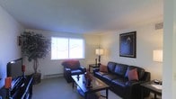 This single-floor space includes a living room that serves as its central gathering area. The room is furnished with a dark leather sofa and a matching armchair, both accented with red and orange throw pillows that add warmth to the otherwise neutral color palette. A rectangular coffee table in a dark finish anchors the seating arrangement, styled with decorative vases and objects. A table lamp with a classic shade provides ambient lighting alongside a side table positioned between the seating pieces. Natural light enters the room through a wide horizontal window, which also offers a partial view of the surrounding neighborhood rooftops. The walls are painted in a soft neutral tone, complemented by a textured ceiling finish. A large framed artwork featuring a dramatic figure portrait adds a bold focal point to one wall, while a decorative dried floral arrangement provides an organic contrast on the opposite side. Baseboard heating units run along the lower walls, indicating a practical heating setup. The flooring transitions subtly between the living area and adjacent spaces, contributing to the overall flow of the layout.
