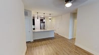 This single-floor space showcases a thoughtfully designed open-concept layout that connects the kitchen and living areas into one cohesive, airy environment. The kitchen serves as a central focal point, anchored by a sleek island with a light-toned countertop that provides both prep space and casual seating. Three pendant lights hang above the island, adding a modern, stylish touch to the space. Dark cabinetry lines the kitchen walls, offering strong contrast against the light countertops, while a stainless steel refrigerator contributes to the contemporary aesthetic. Recessed lighting supplements the pendant fixtures overhead.

Throughout the space, warm light wood-style plank flooring runs continuously, lending a clean and cohesive feel underfoot. Crisp white walls and white-paneled doors maintain a bright, neutral palette that enhances the sense of openness. A ceiling fan is positioned in the living area, adding both functionality and a subtle design element. Multiple doorways are visible throughout, suggesting well-distributed access to additional rooms. The overall ambiance is modern, clean, and move-in ready, with a practical layout suited for everyday living.