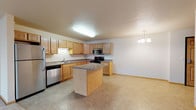 Located at 3400 11th Street SE in Minot, North Dakota, Unit 209 is a thoughtfully designed single-floor residence that offers a functional and comfortable layout. The kitchen serves as a central highlight, featuring warm oak cabinetry with ample storage, laminate countertops, a center island for additional prep and dining space, and stainless steel appliances including a refrigerator, gas range, built-in microwave, and dishwasher. Tile flooring runs throughout the kitchen, which is complemented by a stylish chandelier that transitions the space toward the open living area. The living room is generously sized with plush carpeting and a sliding glass door that opens to a private balcony, allowing for natural light and outdoor access. Bedrooms are carpeted and bright, with windows offering views of the surrounding area and baseboard heating for year-round comfort. The bathroom is clean and functional, featuring a full tub-shower combination, a vanity with oak cabinetry, and tile flooring. A dedicated in-unit laundry area accommodates a full-sized washer and dryer with wire shelving above. Throughout the residence, neutral tones and consistent finishes create a cohesive and inviting atmosphere.
