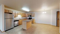 Located at 3400 11th Street SE in Minot, North Dakota, Unit 209 is a thoughtfully designed single-floor residence that offers a functional and comfortable layout. The kitchen serves as a central highlight, featuring warm oak cabinetry with ample storage, laminate countertops, a center island for additional prep and dining space, and stainless steel appliances including a refrigerator, gas range, built-in microwave, and dishwasher. Tile flooring runs throughout the kitchen, which is complemented by a stylish chandelier that transitions the space toward the open living area. The living room is generously sized with plush carpeting and a sliding glass door that opens to a private balcony, allowing for natural light and outdoor access. Bedrooms are carpeted and bright, with windows offering views of the surrounding area and baseboard heating for year-round comfort. The bathroom is clean and functional, featuring a full tub-shower combination, a vanity with oak cabinetry, and tile flooring. A dedicated in-unit laundry area accommodates a full-sized washer and dryer with wire shelving above. Throughout the residence, neutral tones and consistent finishes create a cohesive and inviting atmosphere.