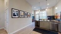 This single-floor residence offers a thoughtfully designed open-concept layout that seamlessly connects the living, dining, and kitchen areas. The space is distinguished by its soaring ceilings, which lend an airy, expansive feel, and light wood-tone flooring that runs throughout the main areas.

The kitchen is outfitted with stainless steel appliances—including a full-size refrigerator, range, and over-the-range microwave—complemented by gray cabinetry, dark countertops, and a white subway tile backsplash. A built-in beverage cooler and a generous breakfast bar with stylish pedestal barstools add both function and visual interest. Pendant lighting illuminates the counter space, enhancing the modern aesthetic.

The living area features large windows and a glass door that flood the space with natural light. A bold navy accent wall anchors the room, creating a striking contrast against the neutral tones elsewhere.

Two bedrooms offer comfortable retreats, each with large windows providing ample daylight. The bathroom presents a dual-sink vanity with dark countertops, gray cabinetry, light wood flooring, and a glass-enclosed shower, delivering a clean, contemporary finish throughout.
