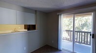 This single-floor space offers a straightforward, functional layout with a comfortable living area finished in neutral tones. The room features wall-to-wall carpeting and smooth-painted walls, creating a clean, understated aesthetic throughout. A textured popcorn ceiling adds a classic touch overhead.

A key feature of the living area is the large sliding glass door, which floods the space with natural light and opens directly onto a private balcony. The balcony overlooks a lush, tree-lined outdoor setting, providing a pleasant natural view and a sense of connection to the surrounding environment.

A pass-through window on one wall connects the living area to the kitchen, offering an open, communicative flow between the two spaces. This design element enhances the sense of openness without fully removing the visual separation between areas. The kitchen area, partially visible through the pass-through, is finished with light-toned countertops.

Overall, the space presents a practical and livable environment with functional layout choices, access to outdoor space, and ample natural light from the sliding glass door.