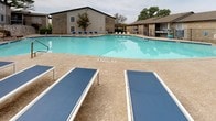 This outdoor amenity space centers around a generously sized swimming pool with clear, blue water, designed for leisure and recreation. The pool area is marked at a maximum depth of 4 feet with a no-diving policy, making it an accessible and family-friendly environment. Surrounding the pool is a broad, textured concrete sun deck finished in a warm sandy tone, providing ample space for relaxation and movement. Blue lounge chairs and chaise lounges are arranged along the pool's edge, offering comfortable spots for sunbathing. A shaded seating area with a table, chairs, and a blue umbrella provides a cooler retreat for those seeking shelter from the sun. The pool deck is bordered by a low-profile natural stone retaining wall, adding a classic, grounded aesthetic to the space. Adjacent to the pool is a single-story clubhouse or leasing office featuring large sliding glass doors, stone column accents, and a covered overhang, blending indoor and outdoor living seamlessly. A gated entrance with black iron fencing provides controlled access to the pool area. The surrounding landscape includes mature trees that offer natural shade and greenery, contributing to a relaxed, resort-like atmosphere within the community.