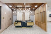Located at 3100 Lake Villa Dr in Metairie, Louisiana, this sophisticated commercial reception space delivers a striking first impression through its carefully curated design elements. The entry is framed by sleek black-trimmed glass double doors, opening onto light-toned herringbone-patterned tile flooring that flows throughout the space.

The centerpiece of the reception area is a dramatic, multi-station desk finished in black-veined marble and polished gold accents, commanding immediate attention. Suspended above it, an elaborate sculptural chandelier composed of cascading crystal cube pendants adds a layer of glamour and visual depth. Warm wood-toned wall paneling and a vertical slatted wood partition screen complement the natural tones of the flooring, creating a cohesive and inviting atmosphere.

Beyond the reception desk, a comfortable lounge area features curved, contemporary seating in muted mauve and white tones, anchored beneath a secondary chandelier and adjacent to large windows that invite natural light. A wall-mounted display screen adds a functional touch to the lounge.

An adjoining convenience retail area, visible through an open archway, offers refrigerated merchandise displays and shelving. Recessed ceiling lighting throughout ensures a bright, welcoming ambiance. The overall design balances luxury with functionality, projecting a polished and professional environment.