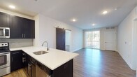Located at 100 Town Center Blvd in Sewell, NJ, this single-floor residence offers a thoughtfully designed open-concept layout with modern finishes throughout. The kitchen is a standout feature, equipped with dark espresso cabinetry, crisp white quartz countertops, a subway tile backsplash, and a full suite of stainless steel appliances including a refrigerator, range, and built-in microwave. A large center island with an undermount sink provides additional workspace and flows seamlessly into the living area. Rich hardwood-style flooring extends throughout the main living spaces, complementing the clean white walls and recessed lighting overhead. A distinctive sliding barn door adds a stylish architectural touch, while floor-to-ceiling vertical blinds frame a sliding glass door that floods the living area with natural light and provides outdoor access. The bedroom offers generous proportions with the same warm flooring and direct access to a well-appointed bathroom. Two bathrooms are present — one featuring a tiled shower-tub combination, and another with a glass-enclosed walk-in shower with decorative mosaic tile accents and modern chrome fixtures. The overall ambiance is bright, clean, and contemporary, making this a well-appointed and move-in-ready residence.