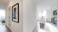 Located at 150 Main St in Hackensack, NJ, this modern single-floor residence offers a thoughtfully designed open-concept layout with clean lines and contemporary finishes throughout. The entryway welcomes you with warm wood-look flooring, a built-in closet, and curated wall art that sets the tone for the rest of the unit. A wide hallway leads into the main living area, where floor-to-ceiling windows flood the space with natural light and frame sweeping views of the surrounding Hackensack skyline. The living room is bright and airy, providing a comfortable and stylish setting for everyday living. The kitchen is a standout feature, equipped with dark cabinetry, stainless steel appliances including a refrigerator, microwave, range, and dishwasher, complemented by a white subway tile backsplash and a central island with a clean white countertop. The bedroom is serene and well-lit, with large windows offering outdoor views and ample space for rest. The bathroom is finished with marble-style tile flooring, white subway tile walls, a dark vanity with a white countertop, and a full soaking tub. Throughout the unit, wood-look flooring, neutral white walls, and modern fixtures create a cohesive, sophisticated aesthetic.