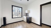 Located on the second floor at 5849 Sunset Boulevard in the heart of Hollywood, Los Angeles, this one-bedroom, one-bathroom unit offers an efficient and thoughtfully designed layout. The main living area features light wood-tone laminate flooring and crisp white walls accented by bold black door frames and baseboards, creating a clean, modern contrast throughout.

A built-in kitchenette occupies one corner of the main room, equipped with white shaker-style cabinetry, a dark countertop, a mosaic tile backsplash in black and white tones, and a compact two-door refrigerator. A large window with horizontal blinds allows natural light to filter in while offering views of the surrounding urban skyline.

The bedroom is accessed through a distinctive black-trimmed doorway and provides a quiet, private retreat with the same consistent flooring and neutral palette. A ceiling fan adds functionality to the space.

The bathroom features a full bathtub with a classic subway-style tile surround in a soft marble-look finish, lending a timeless, clean aesthetic. The unit's design emphasizes simplicity and practicality, making it well-suited for those seeking a low-maintenance residence in one of Los Angeles' most iconic and central neighborhoods.