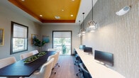 This single-floor business center is a thoughtfully designed workspace that seamlessly blends functionality with contemporary style. The space is divided into two primary zones: a conference area and a dedicated computer workstation lounge, both unified by a striking tray ceiling finished in a bold amber-orange hue that adds warmth and visual interest against the neutral gray walls.

The conference area is anchored by a large, dark wood table surrounded by white upholstered rolling chairs, providing ample seating for meetings and collaborative sessions. Multiple windows with roller shades flood the room with natural light and offer views of the surrounding exterior. Gold-framed abstract artwork adorns the walls, adding a refined aesthetic touch, while a wall-mounted flat-screen display serves as a focal point for presentations.

The workstation lounge features a long, continuous white countertop lined with desktop computers and ergonomic blue task chairs, ideal for individual productivity. A textured accent wall adds depth and sophistication to this area, while a series of geometric glass pendant lights hang overhead, providing both ambient and task lighting. Lush indoor plants are thoughtfully placed throughout, contributing to a fresh, inviting atmosphere. Large windows and a sliding glass door offer outdoor views, further enhancing the bright and open character of the space.