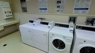 This self-service laundry facility is a practical, single-floor space designed to meet the everyday laundry needs of its users. The room is laid out efficiently, housing a set of commercial-grade Speed Queen washing machines and dryers — including both top-load and front-load washer options — arranged in a clean, accessible row. The machines are equipped with digital control panels and accept credit or debit card payments, providing a modern and cashless user experience. Instructional signage is prominently posted on the wall, guiding users through the step-by-step process of selecting a laundry cycle, inserting payment, and starting the machines. Additional notices and informational sheets are displayed on a bulletin board, ensuring users have all the information they need at a glance. The space features light-colored walls and flooring that contribute to a clean, well-maintained appearance. The layout is straightforward and functional, prioritizing ease of use and efficiency. Overall, this laundry facility offers a no-frills, well-organized environment with reliable commercial equipment suited for regular, high-volume use.