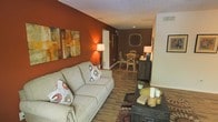 This single-floor space exudes a warm, welcoming ambiance through a carefully curated palette of rich earth tones and thoughtful design details. The living area serves as the heart of the space, anchored by a comfortable beige sofa accented with decorative pillows in complementary rust and cream tones. A bold terracotta accent wall provides a striking backdrop, adorned with a set of abstract canvas art pieces in warm amber and brown hues, adding visual depth and character to the room. Hardwood-style flooring runs throughout, lending a cohesive and grounded feel to the open layout. A dark wood coffee table styled with candles and a woven tray adds a touch of rustic elegance to the living area. The space flows seamlessly into a casual dining area, where a round wooden table surrounded by wicker-style chairs creates an intimate setting for meals. A deep chocolate-brown accent wall featuring a decorative medallion motif anchors the dining space, complemented by soft lamplight that enhances the cozy atmosphere. A dark sideboard provides practical storage while contributing to the space's earthy, layered aesthetic. Overall, the design is cohesive, warm, and richly detailed.