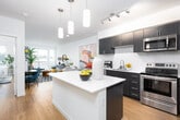 Located at Westminster Way N in Shoreline, Washington, this single-floor residence showcases a contemporary design with an open-concept layout that seamlessly connects the kitchen, dining, and living areas. The kitchen is a standout feature, equipped with dark cabinetry, stainless steel appliances including a refrigerator, range, and microwave, complemented by crisp white quartz countertops and a white subway tile backsplash. A central island with pendant lighting above serves as both a prep space and a visual anchor for the room. Warm hardwood-style flooring runs throughout the main living areas, enhancing the modern aesthetic. The dining area comfortably accommodates a round table and is positioned adjacent to the kitchen for easy flow. The living room opens onto a private balcony through sliding glass doors, offering views of surrounding greenery. The bedroom features a large window that frames outdoor scenery and provides ample natural light. The bathroom presents a clean, minimalist design with a white countertop vanity, undermount sink, and a large mirror. Overall, the space maintains a bright, contemporary ambiance throughout.