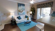 This single-story space centers around a well-appointed living area designed with both comfort and style in mind. The room features rich hardwood flooring throughout, providing a warm and grounded aesthetic. A ceiling fan with an integrated light fixture anchors the space from above, contributing to both airflow and ambient lighting.

The living area is furnished with a plush white sofa accented by teal and blue throw pillows, positioned atop a vibrant turquoise area rug that adds a bold pop of color. A tiered glass-and-chrome coffee table serves as a functional centerpiece, complemented by a matching glass side table nearby. A large abstract painting in shades of blue, white, and gold adorns the wall, reinforcing the room's cohesive color palette.

Natural light flows generously into the space through sliding glass doors dressed with patterned roman shades, offering a view of and access to an outdoor area. A tall indoor plant positioned near the doors introduces a refreshing natural element. Crown molding adds a subtle architectural detail, lending the space a polished, finished character throughout.