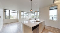 This single-floor unit showcases a clean, contemporary design throughout. The open-concept kitchen serves as a central highlight, featuring a large island with a white quartz countertop, an integrated sink, and a built-in dishwasher. Pendant lights hang above the island, complementing the track lighting overhead. White cabinetry lines the kitchen walls, paired with stainless steel appliances including a refrigerator, range, and microwave. Light wood-tone flooring flows through the kitchen and into the living area, which benefits from an impressive bank of floor-to-ceiling windows offering urban views and abundant natural light.

The bedrooms are carpeted in neutral tones and feature large windows that frame outdoor views of the surrounding neighborhood. Each bedroom provides a quiet, comfortable retreat with clean white walls and ample floor space.

Two bathrooms are present, each featuring a tub and shower combination equipped with accessibility grab bars, light wood-tone flooring, and streamlined vanities with light countertops and ample drawer storage. The overall aesthetic throughout the unit is modern and minimalist, with a neutral color palette of white and warm beige that enhances the sense of openness and light.