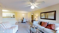 This single-floor residence showcases an open-concept layout that seamlessly connects the living and dining areas, creating a spacious and welcoming atmosphere. The living area is anchored by a plush sofa adorned with decorative pillows in warm earth tones, complemented by a glass-top coffee table that adds a contemporary touch. A ceiling fan with an integrated light fixture serves as a practical focal point overhead, ensuring year-round comfort. Soft, neutral-toned carpeting extends throughout, contributing to the overall warmth of the space.

Adjacent to the living area, the dining space accommodates a round table surrounded by elegant chairs, lit by a classic chandelier that adds a refined ambiance. The warm yellow-toned walls create a cohesive, inviting palette throughout both areas. A large decorative mirror on the living room wall enhances the sense of depth and light. Doorways visible from the main living space suggest access to additional rooms, including what appears to be a bathroom or hallway. The open sightlines between rooms promote an easy flow, making the space both functional and comfortable for everyday living.