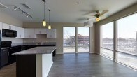 This contemporary single-floor space showcases a thoughtfully designed open-concept layout that seamlessly connects the kitchen and living areas. The kitchen is appointed with sleek white cabinetry, a dark granite countertop island, and pendant lighting with a charming mason jar aesthetic, adding a touch of character to the modern design. A dedicated sink and ample cabinet storage make the kitchen both functional and visually appealing.

The living area is generously proportioned, featuring rich, dark hardwood-style flooring that runs throughout the space, providing a warm and cohesive aesthetic. A ceiling fan with a light fixture ensures year-round comfort. Neutral warm-toned walls complement the flooring and create a welcoming ambiance throughout.

One of the most striking features of this space is the abundance of natural light, courtesy of large sliding glass doors and expansive windows that frame open urban views. The sliding doors provide direct access to the outdoors while flooding the interior with daylight. The views extend to the surrounding cityscape, adding a dynamic, connected feel to the living environment. Overall, the space balances modern finishes with practical design for comfortable urban living.