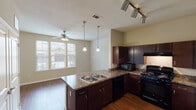 Located at 1200 E Parmer Lane in Austin, TX, this single-floor residence offers a well-designed open layout with consistent hardwood flooring throughout. The kitchen serves as a central feature of the space, boasting dark espresso-toned cabinetry, granite-style countertops, a double-basin sink set into a peninsula island, black appliances including a gas range, microwave, and dishwasher, and both pendant and track lighting that illuminate the workspace effectively. The kitchen opens directly into the living area, which is brightened by a wide set of windows dressed with horizontal blinds and equipped with a ceiling fan, creating a comfortable and airy atmosphere. A hallway connects the main living areas to the private spaces of the unit, including a bedroom that features its own ceiling fan, neutral-toned walls, and ample natural light from multiple windows. The bathroom includes a full soaking tub and shower combination with white tile surround and chrome fixtures, offering a clean and functional design. Throughout the unit, neutral warm-toned walls complement the rich hardwood floors, lending a cohesive and inviting aesthetic. Ceiling fans are present in multiple rooms, enhancing comfort year-round in the Austin climate.