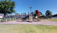 Located at 986 Armfield Circle in Norfolk, Virginia, this single-floor residential community offers a welcoming environment defined by classic brick construction and mature, tree-lined grounds. The exterior grounds are thoughtfully laid out, featuring open green lawn areas, a picnic table for outdoor gatherings, and ample parking for residents — all set beneath the shade of large established trees that lend a peaceful, park-like atmosphere.

The interior common areas are accessed through glass-paneled entry doors that invite natural light into the corridors. The entryway and hallways are finished with durable stone-look tile flooring in warm earth tones, providing a practical yet attractive transition space. Neutral warm-beige walls keep the corridors bright and clean, while framed nautical artwork — depicting sailboats and coastal waterscapes — reinforces the coastal character of the Norfolk setting and adds a refined decorative touch.

The hallways are well-organized, with clearly marked unit doors finished in crisp white paneling and standard safety features, including a fire alarm pull station, ensuring resident safety. A glimpse through the glass entry doors reveals a furnished interior common room with ceiling fans and tile flooring, suggesting comfortable shared amenity spaces within the community. The overall ambiance is tidy, functional, and coastal-inspired.