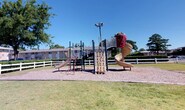 Located at 986 Armfield Circle in Norfolk, Virginia, this single-floor residential community offers a welcoming environment defined by classic brick construction and mature, tree-lined grounds. The exterior grounds are thoughtfully laid out, featuring open green lawn areas, a picnic table for outdoor gatherings, and ample parking for residents — all set beneath the shade of large established trees that lend a peaceful, park-like atmosphere.

The interior common areas are accessed through glass-paneled entry doors that invite natural light into the corridors. The entryway and hallways are finished with durable stone-look tile flooring in warm earth tones, providing a practical yet attractive transition space. Neutral warm-beige walls keep the corridors bright and clean, while framed nautical artwork — depicting sailboats and coastal waterscapes — reinforces the coastal character of the Norfolk setting and adds a refined decorative touch.

The hallways are well-organized, with clearly marked unit doors finished in crisp white paneling and standard safety features, including a fire alarm pull station, ensuring resident safety. A glimpse through the glass entry doors reveals a furnished interior common room with ceiling fans and tile flooring, suggesting comfortable shared amenity spaces within the community. The overall ambiance is tidy, functional, and coastal-inspired.