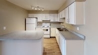 This single-story space is situated on East Tropicana Avenue in Las Vegas, Nevada, offering a practical and well-maintained layout. The kitchen serves as a notable feature of the unit, designed with functionality in mind. It is outfitted with abundant white cabinetry that extends to the upper walls, providing generous storage throughout. Speckled gray countertops run along the perimeter and extend to a peninsula, offering additional workspace and surface area. White appliances — including a refrigerator, electric range, and dishwasher — complement the clean, bright aesthetic of the space. A stainless steel sink is positioned beneath the cabinetry along one wall, adding a practical touch. The kitchen also features wood-look flooring that adds warmth to the overall atmosphere, while a track lighting fixture on the ceiling ensures the area is well-lit. The neutral warm-beige wall tones throughout create a cohesive and inviting ambiance. The single-floor design contributes to ease of movement and accessibility, making it a straightforward and efficient living space in the heart of Las Vegas.