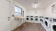 This shared laundry facility is a single-room space designed to serve the practical laundering needs of its users. The room is laid out efficiently, with a row of front-load washing machines lining one wall and multiple stacked dryer units positioned along an adjacent wall, maximizing the available floor area. The machines are coin-operated commercial-grade units, and instructional signage for both washing and drying is posted on the walls for user reference.

The interior features white-painted walls and durable wood-look plank flooring, providing a clean and functional aesthetic. A wall-mounted fold-down table near the window serves as a convenient folding or ironing surface. Natural light enters through a single window on one wall, offering a view of the surrounding outdoor area. Overhead fluorescent lighting ensures the space remains well-lit at all times.

Ventilation grilles are visible near the ceiling, supporting proper airflow within the room. A large utility bin is present for waste disposal. The room is accessed through a standard hinged door. Operating hours are posted near the window, indicating scheduled availability for users. Overall, the space is a straightforward, no-frills laundry facility designed for practicality and ease of use.