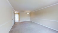 This two-floor property showcases a clean and classic interior design defined by warm neutral tones and timeless architectural detailing. The main living area is generously proportioned, featuring plush wall-to-wall carpeting in a soft grey hue that adds comfort underfoot. Crown moulding and chair rail moulding line the walls, lending the space a refined, traditional character. A delicate chandelier serves as a central focal point, providing ambient lighting that complements the room's understated elegance. The walls are finished in a warm beige palette, creating a welcoming and cohesive atmosphere throughout. Natural light filters into the space through a window visible toward the adjoining area, brightening the interior and enhancing the sense of openness. The layout flows naturally between connected spaces, suggesting a practical and well-considered floor plan. White baseboards and trim provide crisp contrast against the carpeted floors, reinforcing the space's polished, well-maintained appearance. Spanning two floors, this property offers versatile living arrangements suitable for a range of needs.