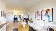 This well-designed single-floor residence offers a thoughtful open-concept layout that seamlessly connects the living, dining, and kitchen areas. Light wood flooring flows throughout the space, complemented by crisp white walls and soaring ceilings that enhance the sense of openness and airiness.

The kitchen is a standout feature, outfitted with flat-front white cabinetry extending to the ceiling, stainless steel appliances including a French door refrigerator, electric range, microwave, and dishwasher, along with a light stone countertop and a kitchen island ideal for casual dining or meal prep.

The living area is bright and welcoming, featuring a ceiling fan and ample room for a full seating arrangement. A dedicated dining space sits just off the kitchen, completing the open social zone.

The bedroom is a serene retreat with large black-framed windows offering views of lush greenery and natural light. It connects to a spacious walk-in closet fitted with wire shelving for generous storage.

The bathroom is clean and modern, featuring a white vanity with stone countertop, a glass-enclosed walk-in shower with white tile surround, and brushed nickel fixtures. The overall aesthetic throughout is contemporary and cohesive, with a neutral palette that creates a calm, functional living environment.