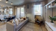 This single-floor residential unit offers a thoughtfully designed layout with a range of functional spaces. The kitchen is outfitted with warm honey-toned wood cabinetry, granite-style countertops, and a full suite of black appliances including a refrigerator, electric range, and over-the-range microwave. Stone-patterned tile flooring runs through the kitchen, which connects directly to a dedicated in-unit laundry area. The living room is bright and welcoming, featuring neutral warm-gray walls, large windows dressed with sheer white curtains, and a sliding glass door that opens to a private outdoor patio, allowing ample natural light to fill the space. The unit includes two bedrooms, both finished with plush carpet and equipped with ceiling fans or pendant lighting. Each bedroom features generous closet storage, with the primary bedroom showcasing a sliding barn-style door leading to a walk-in or reach-in closet. The full bathroom is cleanly finished with stone-patterned tile flooring, a tub-shower combination with white tile surround, a dark wood vanity with countertop storage, and open wall shelving for added functionality. The overall ambiance is calm and cohesive, with a neutral color palette and consistent finishes throughout.