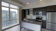 This single-floor space showcases a contemporary design aesthetic with a thoughtful layout and modern finishes throughout. The kitchen serves as a focal point, featuring dark espresso cabinetry that contrasts cleanly against light walls and a neutral tile backsplash. Stainless steel appliances, including a built-in microwave, electric range, and dishwasher, lend a polished, professional quality to the space. A central island with a smooth grey countertop provides additional workspace and casual seating potential. A large window adjacent to the kitchen allows generous natural light to fill the area, offering views of the surrounding urban streetscape. The open layout creates a sense of spaciousness and ease of movement. Recessed and flush-mount ceiling lighting ensures consistent illumination throughout. A dedicated laundry area is conveniently tucked within the unit, equipped with a full-sized stacked washer and dryer set, adding significant everyday practicality. Clean, light-toned flooring runs throughout, complementing the overall neutral and modern palette of the space. Crisp white interior doors with simple panel detailing reinforce the clean, minimalist character of the design.