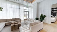 This elegantly designed single-floor amenity space blends contemporary architecture with warm, curated interiors to create a welcoming and visually striking environment. The centerpiece is a grand open lounge area with soaring double-height ceilings, sheer floor-to-ceiling drapery, and expansive glass doors that flood the space with abundant natural light and offer views of the surrounding outdoors. A dramatic linear fireplace set into a marble-clad surround anchors one wall, while a striking sculptural chandelier composed of cascading metallic rods draws the eye upward to a warm wood ceiling accent above. Light hardwood flooring flows throughout, unifying the various zones seamlessly. Generously scaled tufted sofas and sectionals in neutral tones are arranged around rustic wood block coffee tables, creating an inviting social atmosphere. A dedicated media nook features a bold reclaimed wood accent wall housing a flat-screen television, adding an artistic industrial contrast to the otherwise soft palette. The dining area accommodates a large group around a rich wood table paired with iconic modern white chairs, complemented by vibrant large-scale artwork and a sculptural branch chandelier. A refined lounge alcove with built-in dark shelving displays curated books and decorative objects, providing a quieter retreat. Wide corridors with gallery-style lighting connect the areas, with a dedicated mail room accessible along the way.
