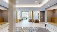 Located at 1710 N Congress Ave in West Palm Beach, FL, this contemporary community amenity space offers a generously sized, open-plan layout spanning a single floor. The design is defined by soaring double-height ceilings at its brightest point, where expansive floor-to-ceiling glass walls flood the interior with abundant natural light and provide sweeping views of the surrounding West Palm Beach streetscape, complete with tropical palm trees.

The space is thoughtfully organized into several distinct functional zones. A lounge area anchored by a sleek billiards table sits beneath a statement pendant light fixture, framed by large-format light tile flooring and bold geometric-patterned area rugs. Adjacent to this is a stylish bar-height counter with seating, ideal for casual dining or co-working. Built-in open shelving and a fluted wood accent wall add warmth and texture to a dedicated lounge nook, complemented by a polished stone feature wall displaying curated artwork.

Recessed banquette seating upholstered in rich golden-amber tones lines a quieter lounge zone, paired with individual side tables and modern floor lamps — creating an inviting setting for relaxed gatherings or focused work. Tray ceilings with integrated ambient lighting enhance the refined, upscale atmosphere throughout the space, making it a versatile and visually striking amenity.
