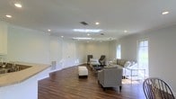 Located at 3975 I-55 N in Jackson, Mississippi, this single-floor space offers a clean, modern aesthetic with a thoughtfully designed open layout. The main living area is expansive and inviting, anchored by rich, dark hardwood flooring that runs throughout and provides a warm contrast to the crisp white walls and trim. Recessed lighting is installed across the ceiling in a deliberate arrangement, ensuring even, ambient illumination throughout the space. A notable skylight feature adds an additional source of natural light, brightening the interior and creating an airy atmosphere. Crown molding along the upper edges of the walls adds a refined architectural detail. The open floor plan allows for flexible furniture arrangements, as demonstrated by the generously sized seating area that accommodates multiple conversation zones. A raised platform or built-in counter element along one wall offers functional versatility. Windows along the perimeter invite natural light and views of the surrounding exterior. The overall ambiance is polished and neutral, making the space highly adaptable for a variety of uses. The combination of contemporary finishes, thoughtful lighting design, and open, flowing layout makes this a well-appointed and functional property.