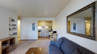 This single-floor residence offers a practical and comfortable open-concept layout that seamlessly connects the main living area to the kitchen. The living room features wall-to-wall carpeting and is furnished with a plush blue sofa and a wooden coffee table, creating a relaxed and inviting atmosphere. A large ornate-framed mirror on the wall adds a decorative touch and enhances the sense of space. The vaulted ceiling rises above the living area, contributing to an airy, open feel throughout. The kitchen is accessible directly from the living space and is outfitted with light wood cabinetry providing ample storage, a gas range, and under-counter appliances. A compact dining nook with a wooden chair and small desk area sits at the transition between the two spaces, making efficient use of the open floor plan. The kitchen features hardwood-style flooring that contrasts neatly with the carpeted living area. Neutral wall tones and warm lighting throughout the space create a cohesive, understated aesthetic. The overall layout prioritizes functionality and ease of movement, making this a well-organized and livable single-floor home.