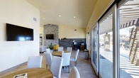 Located at 2100 N 10 in El Centro, this single-story community space offers a welcoming and well-appointed gathering area designed with modern aesthetics in mind. The main lounge is a standout feature, showcasing a striking stacked-stone fireplace that rises to the ceiling and serves as the room's focal point, paired with a wall-mounted flat-screen TV for entertainment. Warm yellow walls contrast elegantly with wood-look tile flooring, creating a bright and inviting atmosphere throughout. Recessed lighting illuminates the space evenly, complementing the natural light that pours in through large sliding glass doors along one wall, which also provide seamless access to an outdoor area. The interior is thoughtfully furnished with round wood dining tables, crisp white upholstered chairs, and accent seating including vibrant yellow chairs that add a pop of color. A dedicated workstation area along the back wall features a desk, computer setup, and shelving, making the space functional for both social gatherings and individual use. A dark stone accent wall adds depth and texture near the workstation, contributing to the space's contemporary character. Overall, the design balances comfort, utility, and modern style in a well-lit, open-plan layout.