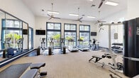 Located at 8585 Woodway Dr in Houston, TX, this fitness center offers a well-appointed, single-level exercise space designed to support a full range of workout routines. The room features notably high ceilings that enhance the sense of openness, while multiple large, navy-framed windows along one wall flood the interior with abundant natural light and provide views of the surrounding greenery. Three ceiling fans are suspended overhead, promoting consistent airflow throughout the space.

The cardio section is well-stocked with treadmills, elliptical trainers, stationary bikes, and a rowing machine, all positioned to take advantage of the natural light from the windows. A multi-station cable machine anchors the strength-training area, complemented by a dumbbell rack, adjustable benches, and additional cable attachment equipment mounted along the walls. Wall-mounted flat-screen televisions are positioned on opposing walls for convenient viewing during workouts.

Neutral-toned carpet tiles cover the flooring throughout, while large framed mirrors on one wall visually expand the space and allow users to monitor their form. Navy blue accent trim along the baseboards and door frames ties the design together, giving the facility a clean, modern aesthetic. Blue-framed glass doors provide a transparent entry, maintaining an open and inviting atmosphere.