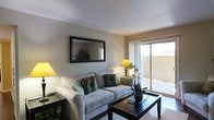 This single-floor residence features a well-appointed living room that serves as the centerpiece of the space. The room is finished with smooth, neutral-toned walls that create a warm and inviting atmosphere. A textured ceiling adds subtle visual interest overhead, while the flooring is covered with a neutral area rug that anchors the seating arrangement.

The living area is furnished with a plush, light gray sofa accented with decorative green and patterned throw pillows, paired with a dark-finished wooden coffee table. A large, dark-framed rectangular mirror is mounted on the main wall, enhancing the sense of depth and reflecting light throughout the room. Ambient lighting is provided by yellow-shaded table lamps positioned on either side of the room, contributing to the warm, comfortable ambiance.

A standout feature of the space is the sliding glass door on one wall, which allows natural light to filter in through sheer white curtains and provides direct access to an outdoor area. This connection to the exterior adds a sense of openness to the interior. Overall, the space presents a clean, cohesive design with a comfortable and functional layout.