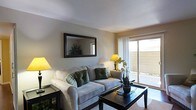 This single-floor residence features a well-appointed living room that serves as the centerpiece of the space. The room is finished with smooth, neutral-toned walls that create a warm and inviting atmosphere. A textured ceiling adds subtle visual interest overhead, while the flooring is covered with a neutral area rug that anchors the seating arrangement.

The living area is furnished with a plush, light gray sofa accented with decorative green and patterned throw pillows, paired with a dark-finished wooden coffee table. A large, dark-framed rectangular mirror is mounted on the main wall, enhancing the sense of depth and reflecting light throughout the room. Ambient lighting is provided by yellow-shaded table lamps positioned on either side of the room, contributing to the warm, comfortable ambiance.

A standout feature of the space is the sliding glass door on one wall, which allows natural light to filter in through sheer white curtains and provides direct access to an outdoor area. This connection to the exterior adds a sense of openness to the interior. Overall, the space presents a clean, cohesive design with a comfortable and functional layout.