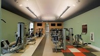 This fitness center is designed to support a comprehensive workout experience within a single-level layout. The space is organized into distinct zones, with cardio equipment such as ellipticals and upright bikes lining one side, while a robust selection of strength-training cable machines and resistance equipment occupies the other. The elongated floor plan allows for easy circulation between stations without crowding. Overhead, rows of long linear pendant lights and recessed fixtures provide bright, even illumination throughout. The walls feature a two-tone color scheme, pairing sage green upper sections with warm orange accents near the center, creating an energizing atmosphere. Multiple flat-screen televisions are mounted along the central wall, keeping members entertained during workouts. The flooring incorporates a durable multi-tone design with checkered accents in red, gray, and neutral tones, adding visual interest underfoot. Natural light enters through windows at one end of the room, contributing to the open and welcoming feel of the space. Overall, the facility is well-maintained and thoughtfully arranged to accommodate a variety of fitness routines.