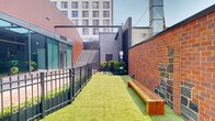 This urban outdoor courtyard is a well-designed ground-level space that blends modern amenities with its surrounding architectural context. The area is laid with lush artificial turf that runs the length of the courtyard, creating a clean, green surface framed by contrasting hard landscaping elements.

On one side, a contemporary building with floor-to-ceiling glazed panels and a distinctive terracotta-red facade provides a strong visual boundary. Through the glass walls, fitness equipment is visible, suggesting an adjacent gym or wellness facility. A set of concrete stairs with metal railings ascends along the building exterior, providing access to upper levels.

The opposite boundary is defined by a heritage-style red brick wall, adding textural contrast and a sense of history to the space. Timber slatted benches are positioned along this wall, offering casual seating for occupants. Black powder-coated steel fencing runs along the courtyard's perimeter, with a gated entry providing controlled access.

At the far end, the courtyard opens toward a lively public plaza featuring colorful street murals, young trees, and planted garden beds, connecting the space visually to the broader urban neighborhood. Security lighting is mounted on posts throughout. The open-air design ensures ample natural light and a pleasant outdoor atmosphere suited for relaxation or pet exercise.