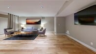 Located at 13900 Tahiti Way in Marina del Rey, California, this thoughtfully designed single-floor space offers a seamless blend of lounge and meeting functionality. Light hardwood flooring in a herringbone-style pattern runs throughout, lending warmth and a sense of openness to the entire layout.

The space is divided into two distinct yet connected zones. The lounge area features a contemporary gray sofa and accent chair arranged around a striking live-edge wood coffee table, anchored by a charcoal area rug. Warm table lamps and a bold panoramic sunset artwork on the wall create an inviting, relaxed ambiance. Built-in open shelving lines one wall, providing additional display and storage space, while a cozy seating nook with vibrant blue accent chairs adds a pop of color.

The conference area is equipped with a large rectangular table surrounded by ergonomic white chairs, ideal for meetings or collaborative work. A wall-mounted flat-screen display faces the table, and a textured area rug grounds the setup. Recessed lighting throughout ensures even, professional illumination across both zones. Warm taupe walls, clean white trim, and curtained room dividers contribute to a cohesive, modern aesthetic that is both functional and welcoming.