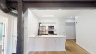 This single-floor unit showcases a compelling blend of industrial heritage and modern design. The open-concept layout is anchored by a sleek kitchen featuring a large peninsula with a marble-veined white countertop, an integrated sink, and white shaker-style cabinetry. Stainless steel appliances — including a refrigerator, microwave, and gas range — are set against a classic subway tile backsplash, completing a clean, functional cooking space. Light hardwood flooring runs throughout, providing warmth and continuity across the unit.

The living area retains striking industrial character with exposed red brick walls, dark structural steel beams traversing the ceiling, and visible ductwork — all hallmarks of a converted loft space. A large double-hung window allows natural light to fill the room, highlighting the texture of the original masonry. Exposed columns add architectural interest and reinforce the unit's industrial roots.

The bedroom offers an open, airy feel with light walls and continued hardwood flooring, while a frosted-glass sliding door provides access to the bathroom. The bathroom is finished with neutral tile flooring and walls, a tub and shower combination enclosed by frosted sliding glass doors, and a vanity with clean, modern fixtures.