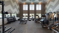 Located at 2015 Wave Crest Dr in Raleigh, NC, this fitness center is designed to accommodate a wide range of workout needs within a single, thoughtfully laid-out floor. The main workout area immediately commands attention with its soaring double-height ceilings and floor-to-ceiling windows that flood the space with natural light and offer views of the surrounding outdoor area. A bold chevron pattern adorns the upper walls, lending visual energy to the room, while carpet flooring provides comfort underfoot throughout. This primary zone is outfitted with multiple treadmills, ellipticals, and a variety of Precor strength machines, including cable systems and resistance equipment, offering users a thorough cardiovascular and strength-training experience. Full-length mirrors and a dedicated dumbbell rack further enhance functionality. An adjoining studio area, finished with neutral tones, crown molding, and recessed lighting, provides a quieter, open space ideal for group exercise, stretching, or functional training. This room features step platforms, resistance accessories, and stability balls stored on wall-mounted racks, along with a ballet barre and ample open floor space. Additional amenities include built-in shelving and storage closets. Together, the spaces create a comprehensive, well-designed fitness facility suited for a broad range of fitness activities.