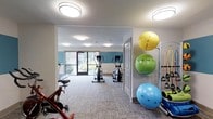 The fitness center is a thoughtfully designed, single-floor facility that provides a clean and welcoming environment for exercise. The space features a neutral color palette with a teal accent wall that adds a pop of color and energy to the room. Flush-mounted circular ceiling lights are distributed evenly throughout, ensuring consistent, bright illumination across the entire area.

The cardio section is well-equipped, housing multiple elliptical machines and a stationary bike, arranged to allow comfortable spacing between users. Large glass doors at the far end of the room flood the space with natural light and provide views of the surrounding greenery, creating an inviting atmosphere.

Along one wall, a vertical rack holds three exercise balls in vibrant yellow, green, and blue, adding both functionality and visual interest. Nearby, resistance bands are mounted and readily accessible. The room's neutral, textured carpet adds warmth underfoot while maintaining a practical surface for varied workout activities.

An adjoining room, accessible through a doorway, extends the usable workout area, offering additional space for equipment or stretching. Overall, the fitness center balances practicality with a bright, modern aesthetic.