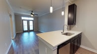Located at 11 Parklands Drive in Bluffton, South Carolina, this single-floor residence offers a thoughtfully designed open-concept layout that seamlessly connects the kitchen and living areas. The kitchen is outfitted with rich dark cabinetry, a clean white countertop peninsula with a stainless steel sink, and elegant pendant lighting that adds a modern touch. A tile backsplash and built-in dishwasher round out the functional and attractive kitchen space. The living area features warm hardwood-style flooring, crown molding, and soft neutral wall tones that create a bright and welcoming atmosphere. A ceiling fan with a light fixture enhances comfort throughout the main living space. French doors at the far end of the living area flood the room with natural light and provide direct access to an outdoor balcony, offering a seamless indoor-outdoor connection. The overall design balances contemporary style with practical living, making this Bluffton property a well-appointed and comfortable space suited for everyday life.