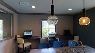 This single-floor space serves as a functional and stylish business center and collaborative meeting area. The room is anchored by exposed brick walls painted in a neutral tone, lending a warm, urban character to the interior. A built-in desk runs along the perimeter wall, providing dedicated workstations equipped with computers and a multifunction printer, making it well-suited for productivity and office tasks.

A central conference-style table surrounded by vibrant blue upholstered chairs creates an inviting gathering area, ideal for meetings or group work. Decorative accents, including a globe-shaped pendant light with an ornate dark fixture, add a touch of character and visual interest to the space.

Natural light flows in through a centrally positioned window that frames a lush, green outdoor view, contributing to a refreshing and pleasant atmosphere. Recessed ceiling lighting supplements the natural light, ensuring the space remains bright and well-illuminated throughout the day.

The overall design balances practicality with aesthetic appeal, combining modern technology and comfortable seating with classic architectural details. This space is well-suited for professionals seeking a productive environment equipped with essential amenities.