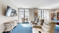 This single-floor amenity lounge offers a well-rounded recreational experience within a compact yet efficiently utilized footprint. The space is designed with a modern aesthetic, featuring white walls, recessed lighting, and a bold blue multi-tone carpet that anchors the room with a contemporary feel.

The lounge is divided into distinct activity zones. A media area is furnished with rows of plush leather recliner seats equipped with cup holders, positioned in front of a wall-mounted flat-screen television — ideal for group viewing or casual relaxation. Adjacent to this, a dedicated games area hosts a full-size billiards table with a blue felt surface, a foosball table, and additional tabletop games, clearly marked with billiard signage overhead.

A snack and beverage station lines one wall, featuring refrigerated drink cases and stocked shelving units, functioning as a self-serve convenience kiosk. A standing-height bar table with stools provides additional seating nearby.

Large floor-to-ceiling windows and a glass sliding door flood the interior with abundant natural light and offer views of an outdoor terrace area. A decorative column adds an architectural accent, while colorful artwork on the walls contributes to the space's lively, social atmosphere.