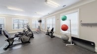 This single-floor amenity space is thoughtfully designed to serve both fitness enthusiasts and families with young children. The facility is divided into two distinct areas connected by an open layout, as visible in the dollhouse overview.

The fitness center occupies the larger portion of the space and is generously outfitted with a wide range of equipment. Cardio machines — including ellipticals, stationary bikes, and treadmills — are arranged across durable rubber flooring, providing ample room to move between stations. Strength training is well represented with multiple cable and weight machines featuring red upholstered seats, a dumbbell rack, and a weight bench. Exercise balls in multiple sizes are neatly stored on a dedicated rack. Multiple windows fitted with horizontal blinds allow natural light to fill the room, while wall-mounted televisions and bright overhead fluorescent lighting enhance the workout environment. Crown molding and neutral beige walls give the space a polished, clean aesthetic.

The children's play area adjoins the gym and is a cheerful, kid-friendly environment. It features a colorful alphabet and U.S. map area rug, a wall-mounted chalkboard, a puppet theater, and a navy-striped play teepee. Whimsical animal decals dot the walls, creating an engaging and imaginative atmosphere for younger visitors.